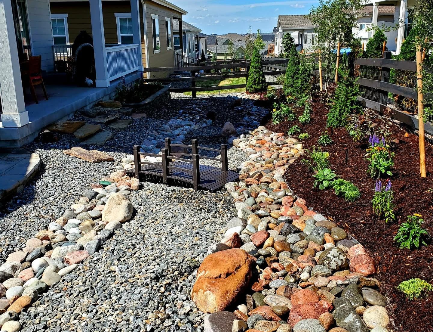 A dry creek bed of river stones with a wooden footbridge, edged by new water-wise plantings and mulch
